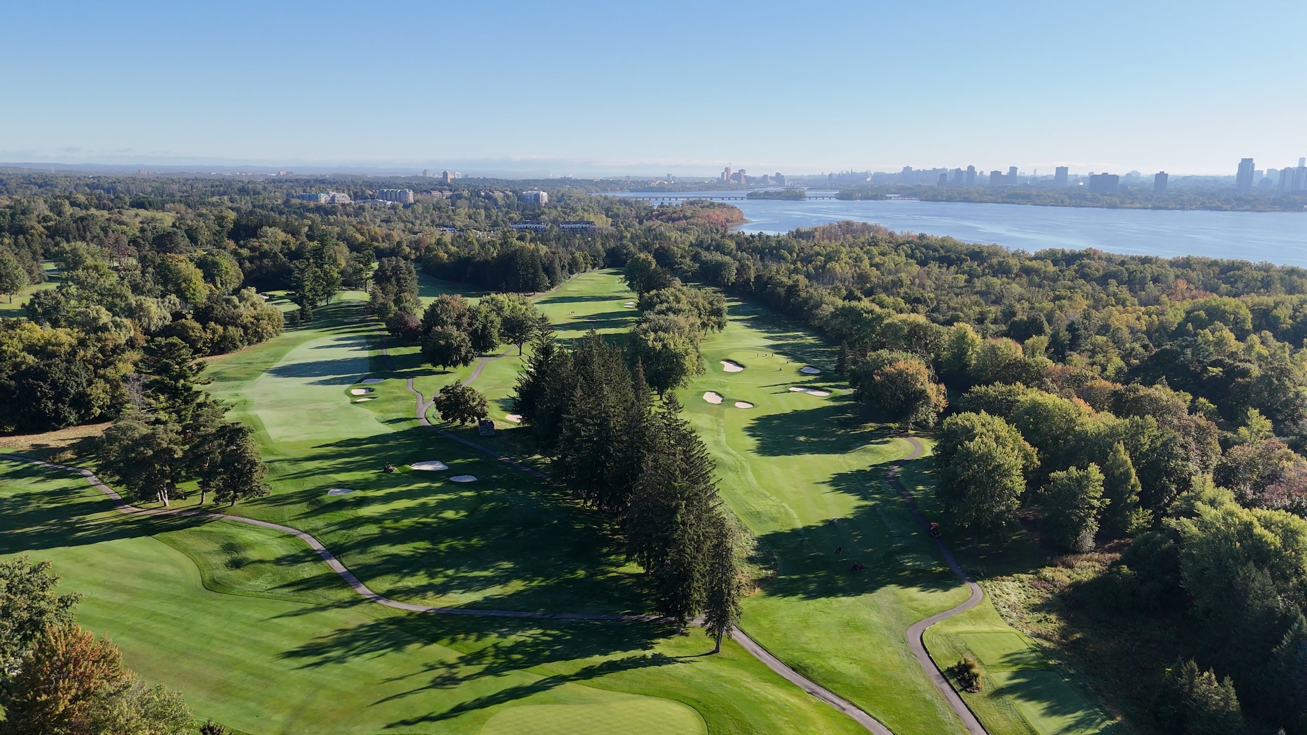 Aerial view of Rivermead's tree-lined fairways beside the Ottawa River, with the Ottawa skyline beyond