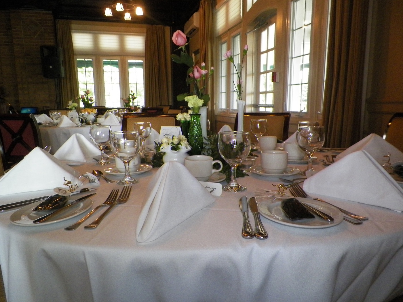Tables in the dining room set with white linens, roses, and folded napkins, tall windows beyond
