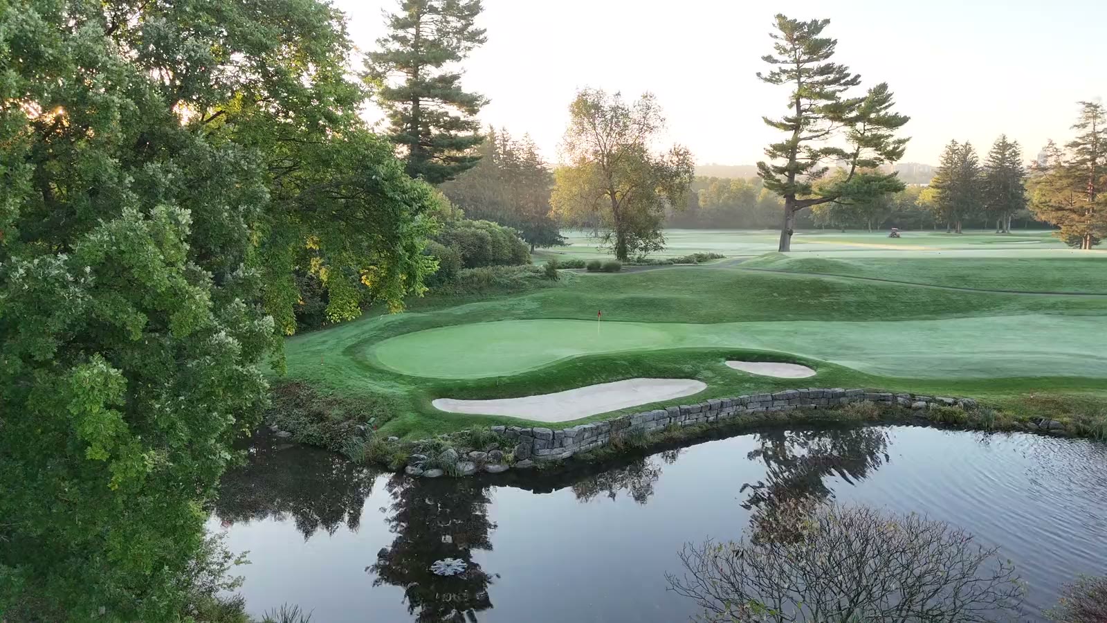 Drone view over a Rivermead green at dawn, with the stone-walled pond in the foreground