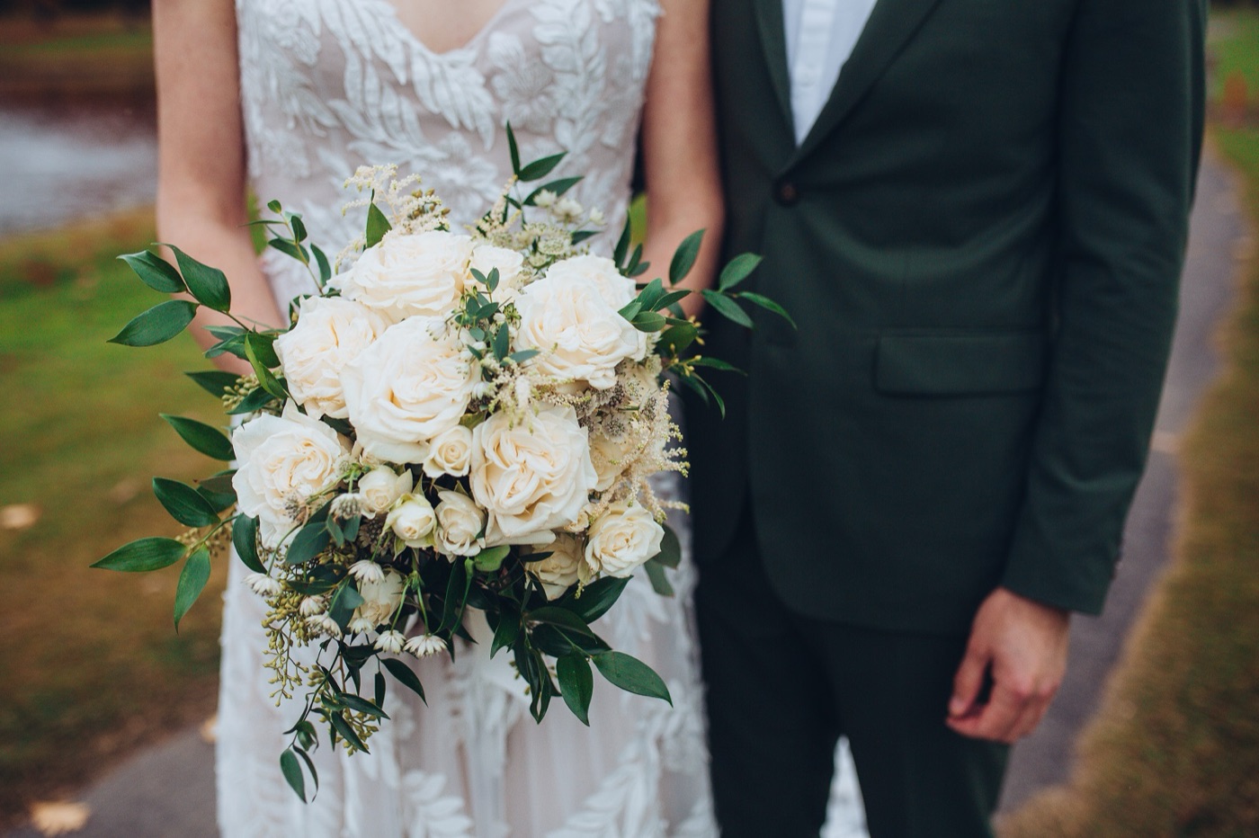 A bouquet of white roses held between the bride and the groom's deep green suit