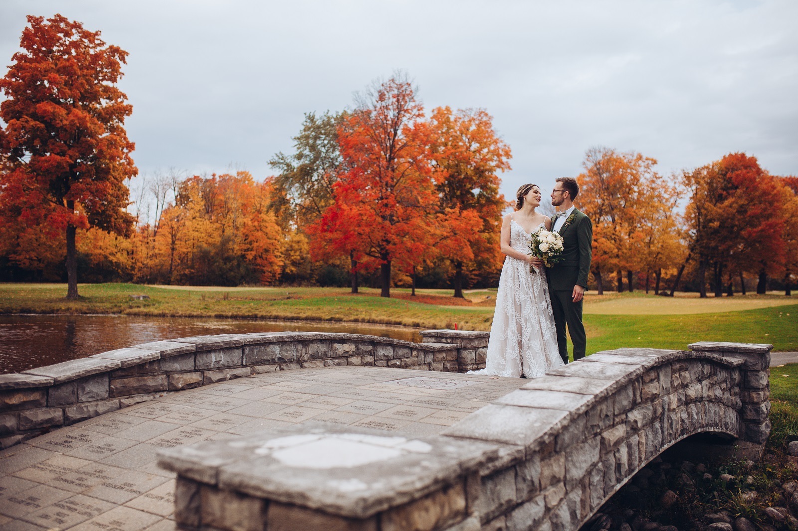 A bride and groom on Rivermead's engraved stone bridge, surrounded by red and orange maples